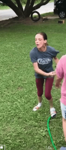 A Woman In A Blue Shirt With The Word Faith On It Is Jumping Over A Green Hose . GIF