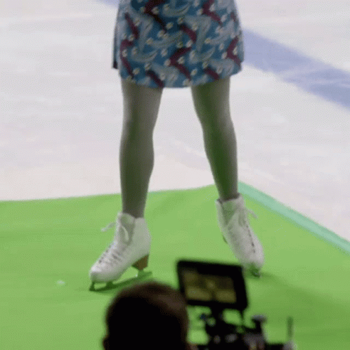 A Woman In A Blue Skirt Is Standing On A Green Mat Wearing Ice Skates GIF