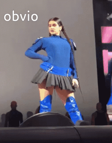 A Woman In A Blue Sweater And Blue Boots Stands On A Stage With The Word Obvio Written Above Her GIF