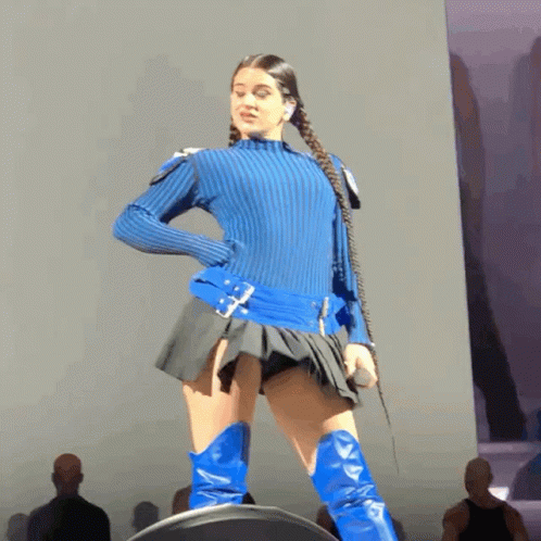 A Woman In A Blue Sweater And Blue Boots Stands On Stage GIF