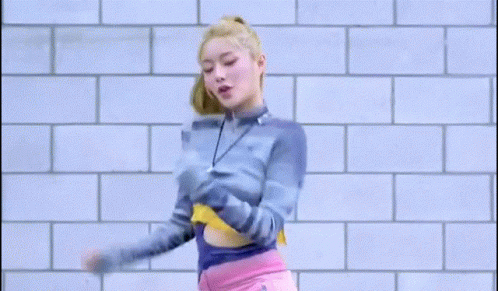 A Woman In A Blue Sweater And Pink Shorts Is Dancing In Front Of A Brick Wall . GIF