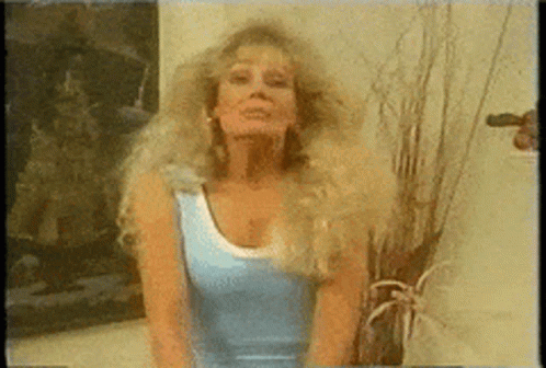A Woman In A Blue Tank Top Stands In Front Of A Plant GIF