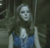 A Woman In A Blue Top And Necklace Is Standing In A Dark Room . GIF