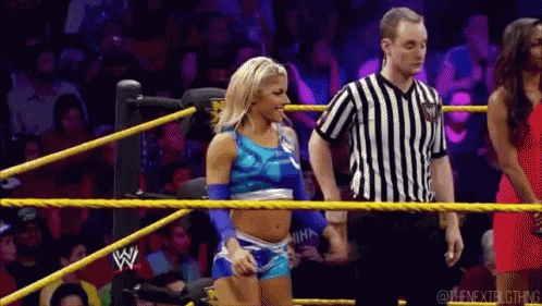 A Woman In A Blue Top Is Standing In A Wrestling Ring Next To A Referee Wearing A Black And White Striped Shirt GIF
