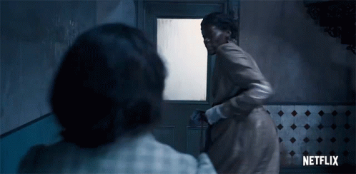 A Woman In A Brown Coat Is Standing In Front Of A Door In A Room . GIF