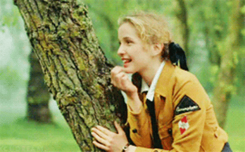 A Woman In A Brown Jacket Is Leaning Against A Tree Trunk And Smiling GIF