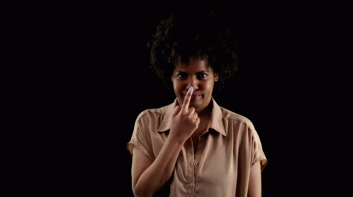 A Woman In A Brown Shirt Holds Her Finger To Her Nose Against A Black Background GIF