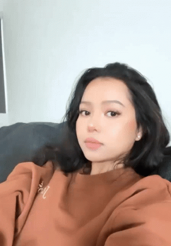A Woman In A Brown Sweatshirt Is Sitting On A Couch Taking A Picture Of Herself . GIF