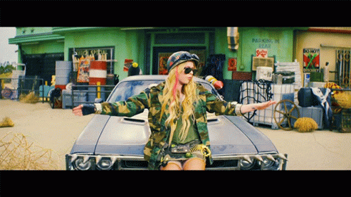 A Woman In A Camouflage Jacket Is Standing Next To A Car In Front Of A Building That Says Parking Is Illegal GIF