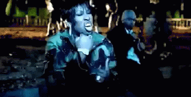 A Woman In A Camouflage Jacket Sings Into A Microphone In Front Of A Man In A Suit GIF