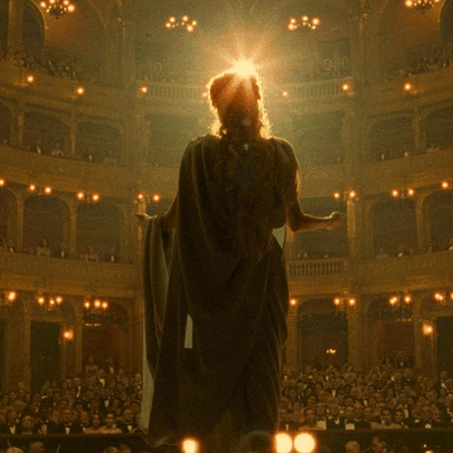 A Woman In A Cape Stands In Front Of A Crowd In A Theater GIF