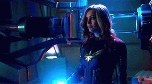 A Woman In A Captain Marvel Costume Is Standing In Front Of A Blue Light GIF