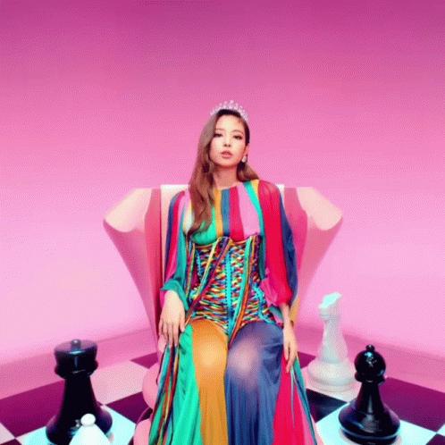 A Woman In A Colorful Dress Is Sitting On A Chair With Chess Pieces Around Her GIF
