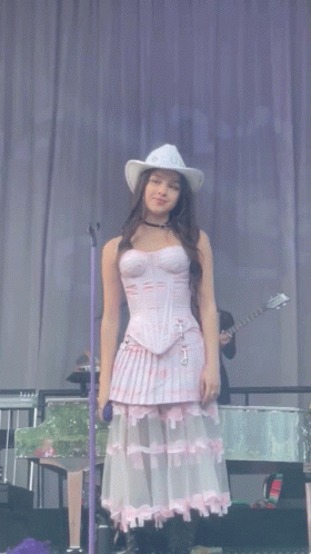 A Woman In A Corset And Cowboy Hat Stands In Front Of A Microphone On Stage GIF