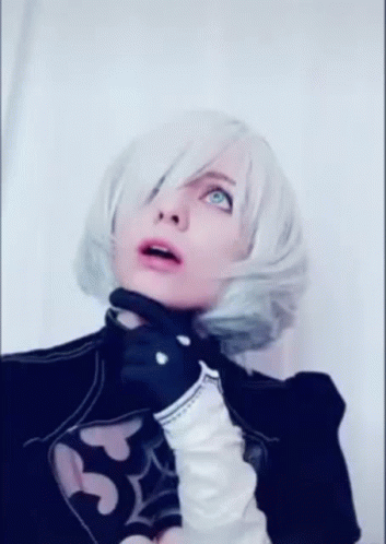 A Woman In A Cosplay Costume With White Hair And Black Gloves Is Standing In Front Of A White Wall . GIF