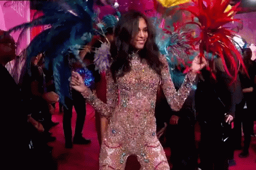 A Woman In A Costume With Feathers On Her Head Is Dancing On A Pink Carpet . GIF