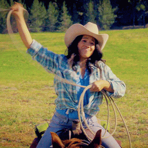 A Woman In A Cowboy Hat Is Holding A Lasso While Riding A Horse GIF