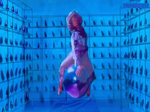 A Woman In A Cowboy Hat Is Riding A Purple Object In A Room With A Lot Of Shoes On Shelves . GIF