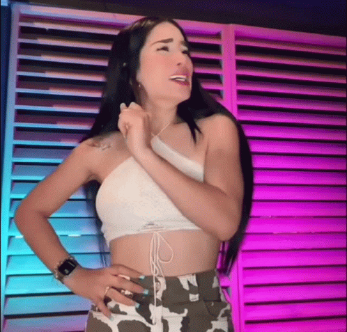 A Woman In A Crop Top And Camo Shorts Is Standing In Front Of A Purple Wall . GIF