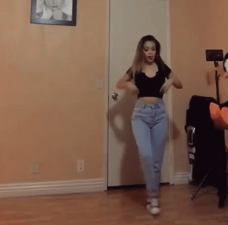 A Woman In A Crop Top And Jeans Is Dancing In A Room . GIF
