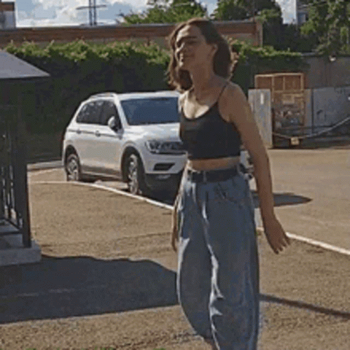 A Woman In A Crop Top And Jeans Is Standing In A Parking Lot . GIF