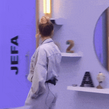 A Woman In A Denim Jacket And Pants Stands In Front Of A Blue Wall GIF