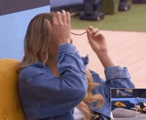 A Woman In A Denim Jacket Is Covering Her Face With Her Hands While Holding A Pair Of Glasses . GIF