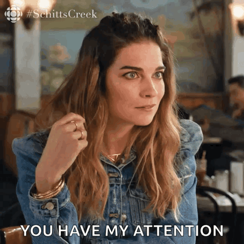 A Woman In A Denim Jacket Is Sitting At A Table And Says " You Have My Attention " GIF