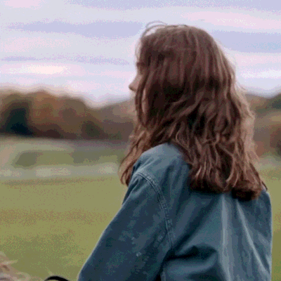 A Woman In A Denim Jacket Is Standing In A Field With Her Back To The Camera GIF