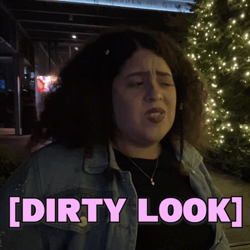 A Woman In A Denim Jacket Is Standing In Front Of A Sign That Says [ Dirty Look ] GIF