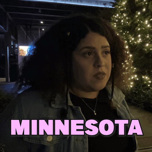 A Woman In A Denim Jacket Is Standing In Front Of A Sign That Says Minnesota GIF
