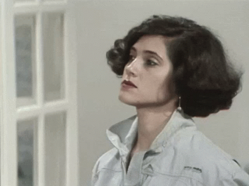 A Woman In A Denim Jacket Is Standing In Front Of A Window And Looking At The Camera . GIF