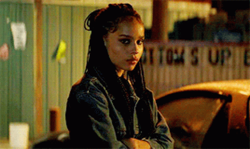 A Woman In A Denim Jacket Is Standing Next To A Car With Her Arms Crossed And Looking At The Camera . GIF