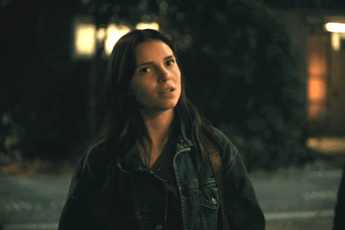 A Woman In A Denim Jacket Stands In Front Of A Building At Night GIF