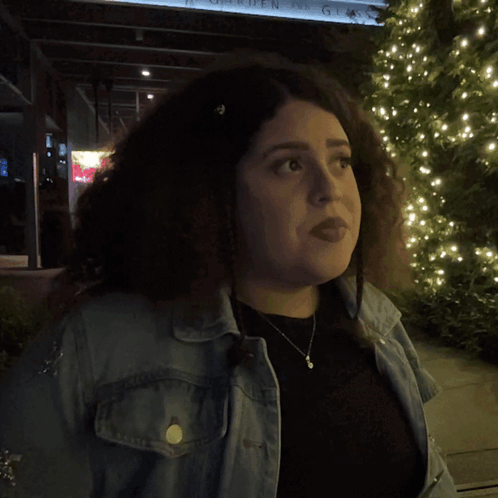 A Woman In A Denim Jacket Stands In Front Of A Sign That Says What GIF
