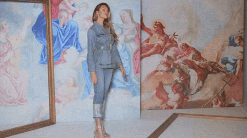 A Woman In A Denim Jacket Stands In Front Of A Wall Of Paintings GIF
