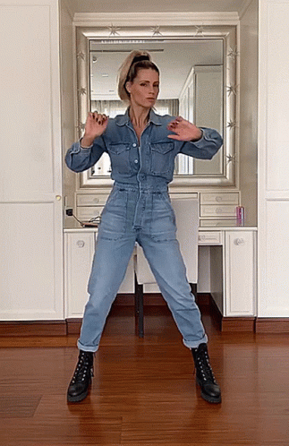 A Woman In A Denim Jumpsuit And Black Boots Is Dancing In Front Of A Mirror GIF