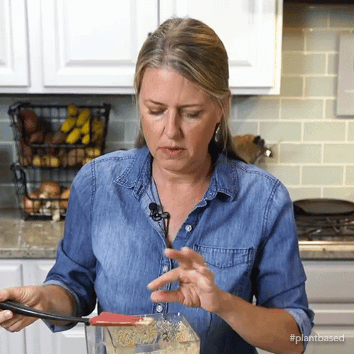 A Woman In A Denim Shirt Is Stirring Something In A Blender With The Hashtag #Plantbased On The Bottom GIF