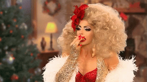A Woman In A Drag Queen Costume Is Sitting In Front Of A Christmas Tree And Eating Something . GIF