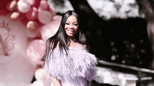 A Woman In A Feathered Dress Is Standing In Front Of Balloons And Smiling . GIF