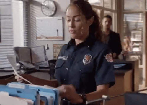 A Woman In A Firefighter 'S Uniform Is Standing In An Office Looking At A Stack Of Papers . GIF