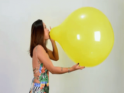 A Woman In A Floral Dress Is Blowing Up A Large Yellow Balloon . GIF