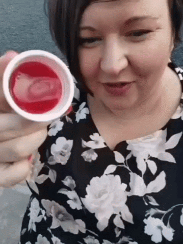 A Woman In A Floral Dress Is Holding A Cup Of Liquid In Her Hand . GIF