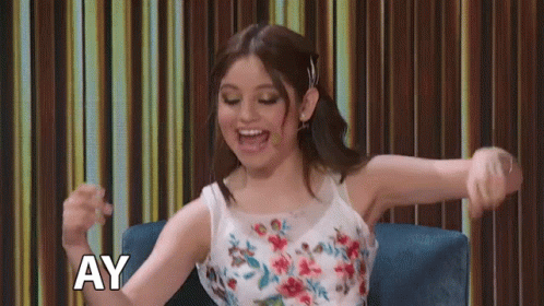 A Woman In A Floral Dress Is Sitting In A Chair With Her Arms Outstretched And The Word Ay Above Her GIF
