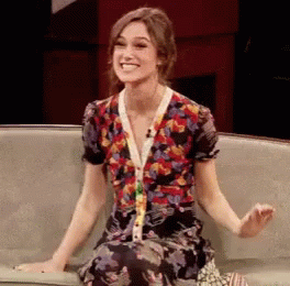 A Woman In A Floral Dress Sits On A Couch Smiling GIF