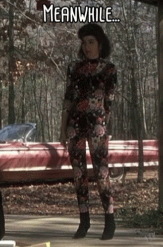 A Woman In A Floral Jumpsuit Is Standing In Front Of A Red Car With The Words Meanwhile Below Her GIF