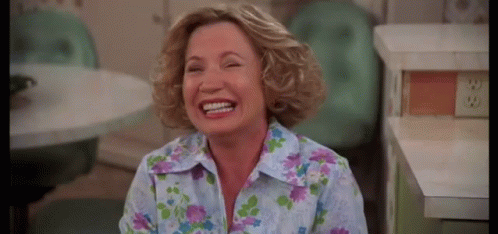 A Woman In A Floral Shirt Is Laughing In A Kitchen While Sitting At A Table . GIF