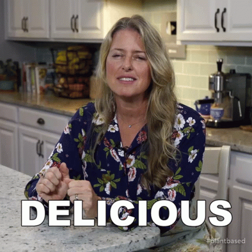 A Woman In A Floral Shirt Is Sitting At A Kitchen Counter And The Word Delicious Is Above Her GIF