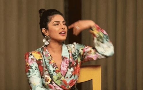 A Woman In A Floral Shirt Is Sitting In A Chair And Pointing At Her Face GIF