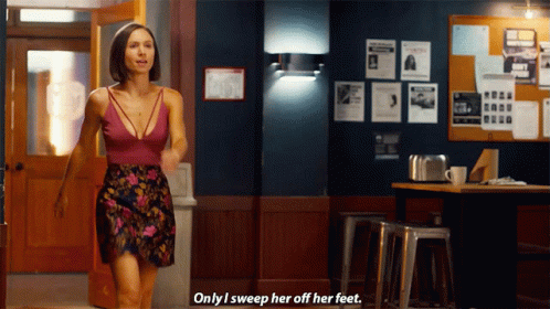 A Woman In A Floral Skirt Says " Only I Sweep Her Off Her Feet " In Front Of A Bulletin Board GIF